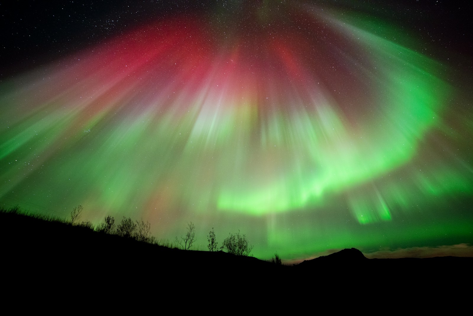 Red and green northern lights filling the night sky over a hillside